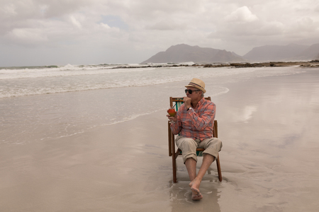 Front view of active senior man having cocktail drink while relaxing in a sun lounger at the beach with moutains and ocean in the background. He wears a hatの写真素材