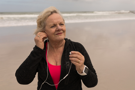 Front view of active senior woman wearing earphones at the beach with ocean in the background. She seems happyの写真素材