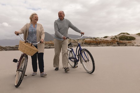 Front view of active senior couple walking with bicycle at the beach with mountains in the background. They seem happyの写真素材