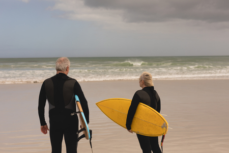 Rear view of senior couple standing with surfboard on the beachの写真素材