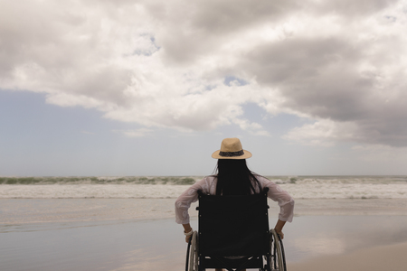 Rear view of disabled young woman with hat sitting on wheelchair at beachの写真素材