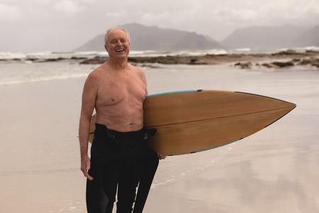 Front view of active senior man standing with surfboard on the beach against mountains background