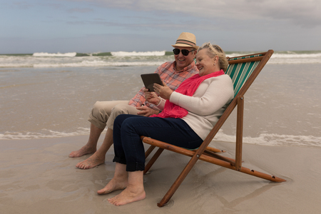 Side view of active senior couple using digital tablet while relaxing in a sun lounger at the beach with ocean in the background. They seem happyの写真素材
