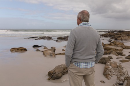 Rear view of active senior man standing with hands in pocket at beachの写真素材