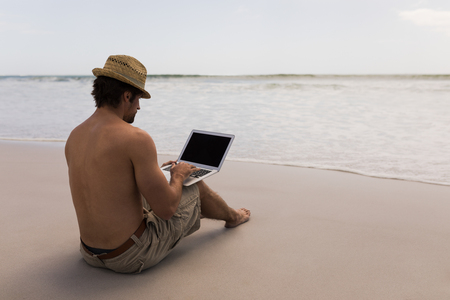 Rear view of shirtless young man with hat using laptop on the beachの写真素材