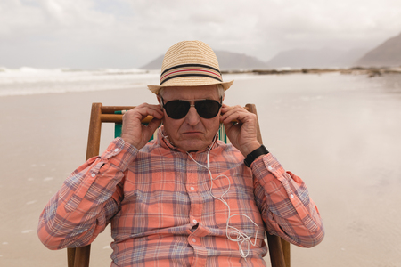 front view of active senior man wearing earphones while relaxing in a sun lounger on the beach. He wears a hatの写真素材