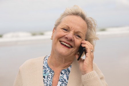 Front view of active senior woman talking on mobile phone standing at beach with ocean in the background. She seems happy