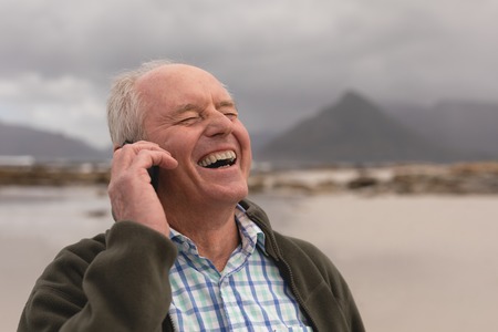Side view of active senior man talking on mobile phone standing at beach with mountains in the background. He seems happyの写真素材