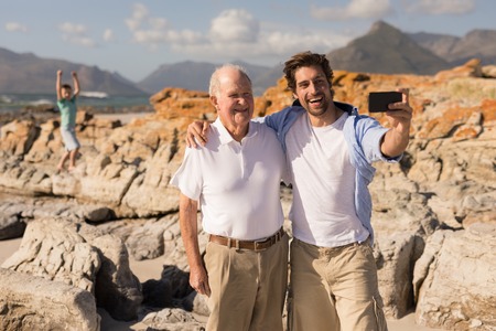 Front view of happy father and son with arms around taking selfie with mobile phone on beach