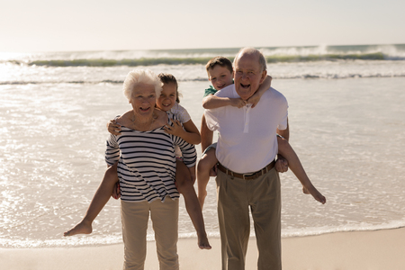 Front view of happy grandparents giving piggyback to grandchildren on beach in the sunshineの写真素材