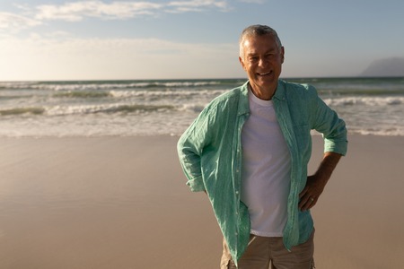 Front view of happy senior man standing with hands on hip at beachの写真素材