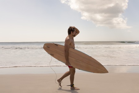 Side view of shirtless young male surfer with surfboard standing on beach in the sunshine