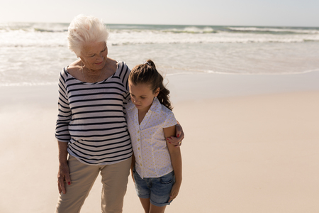 Front view of senior woman and her granddaughter with arms around standing on beach in the sunshineの写真素材