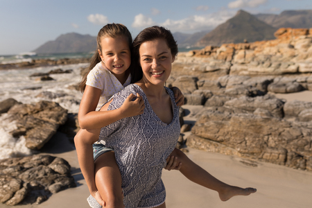Front view of beautiful young mother giving piggyback to her daughter on beachの写真素材