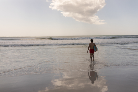 Rear view of shirtless young male surfer with surfboard walking on beachの写真素材