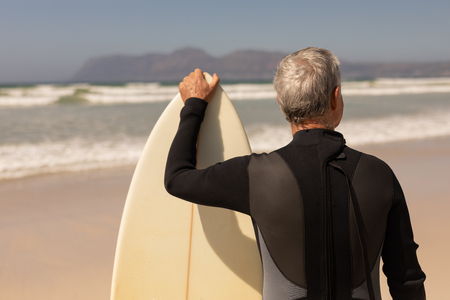 Rear view of senior male surfer standing with surfboard on the beach with mountains in the backgroundの写真素材
