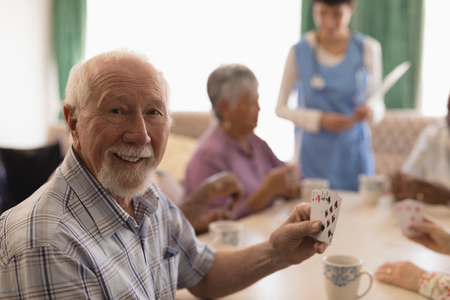 Front view of senior man playing cards with senior people and female doctor on behind in living room...