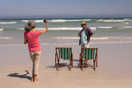 Rear view of senior woman clicking photo of senior man on beach in the sunshineの写真素材