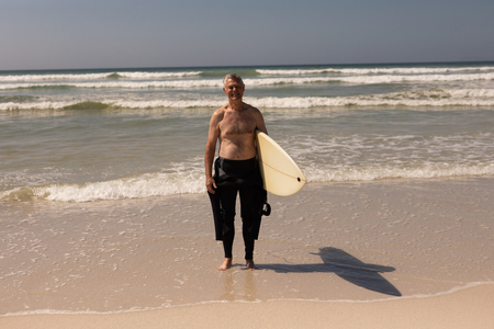 Front view of active senior male surfer standing with surfboard on the beachの写真素材
