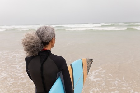 Rear view of senior female surfer walking towards the sea with surfboard on the beach