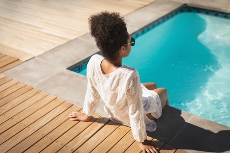 Rear view of young mixed-race woman with sunglasses sitting at poolside in backyard of home on a sunny dayの写真素材