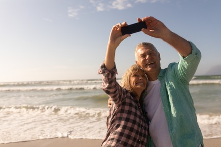 front view of active senior couple taking selfie with mobile phone on the beachの写真素材