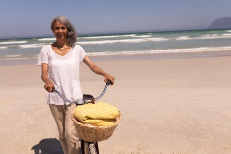 Front view of happy senior woman with bicycle walking on beach in the sunshineの写真素材