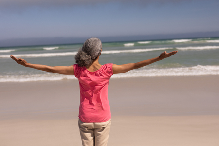Rear view of senior woman with arms stretched out standing on beach in the sunshineの写真素材