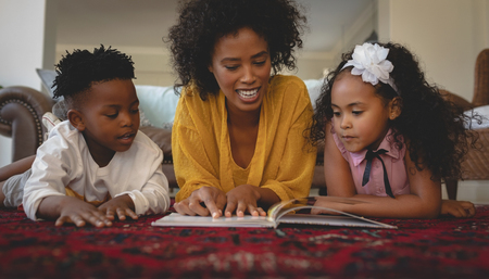 Front view of African American mother with her cute children lying on floor and reading a storybook in a comfortable homeの写真素材