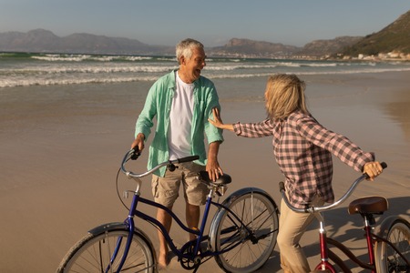 Front view of active senior couple standing with bicycle on the beachの写真素材