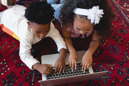High angle view of cute African American sibling lying on floor and using laptop in a comfortable...