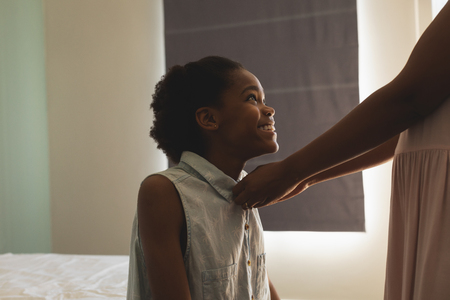 Side view of African American mother helping her daughter to wear a cloth in bedroom at homeの写真素材