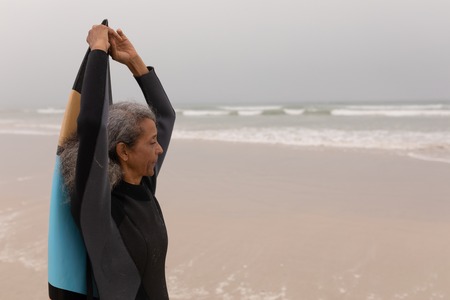 Side view of senior female surfer with arms up holding surfboard on the beach