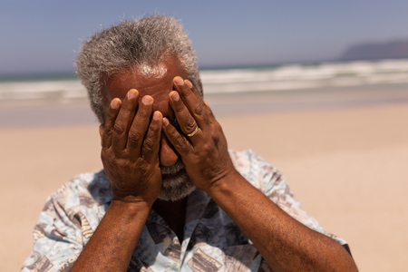 Front view of senior black man with hands on face standing on beach in the sunshineの写真素材