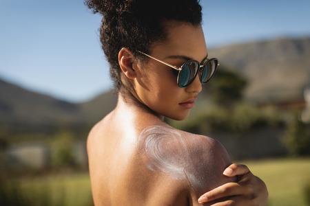 Side view of young mixed-race woman applying sunscreen on shoulders in backyard of home on a sunny...