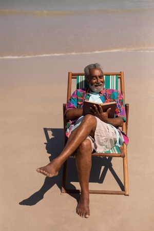 High angle view of senior man relaxing on sun lounger and reading a book on beach in the sunshineの写真素材
