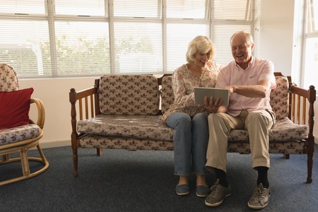Front view of senior couple laughing and using digital tablet in living room at nursing homeの写真素材