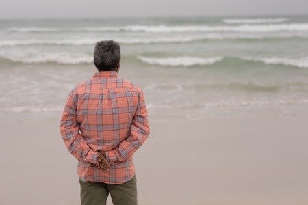 Rear view of senior man with hands behind back standing on the beachの写真素材