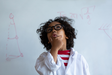Front view of a thoughtful Mixed-race schoolboy in chemistry coat looking above his head while standing in front of the white board in the laboratory at schoolの写真素材