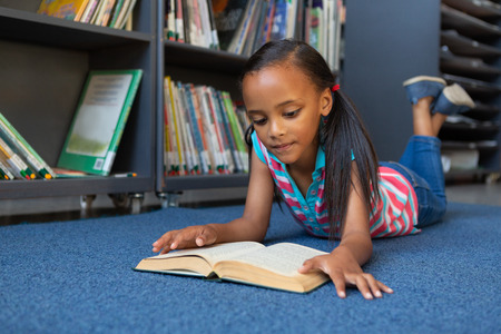 Front view of a cute mixed-race schoolgirl reading a book while lying down in the library at school