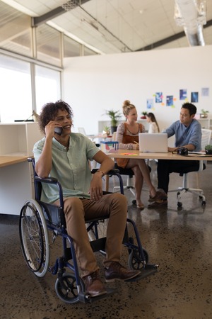 Front view of handsome disabled male executive talking on mobile phone in office with coworkers in the backgroundの写真素材