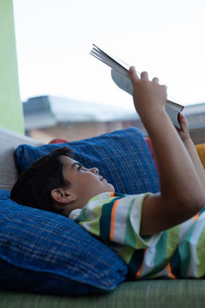Side view of a Caucasian schoolboy lying on sofa and reading a book in library at elementary schoolの写真素材