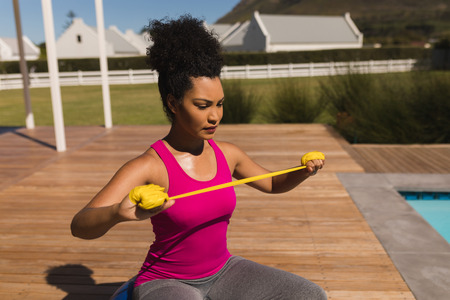 Front view of beautiful young African American woman exercising with resistance band in the backyard...