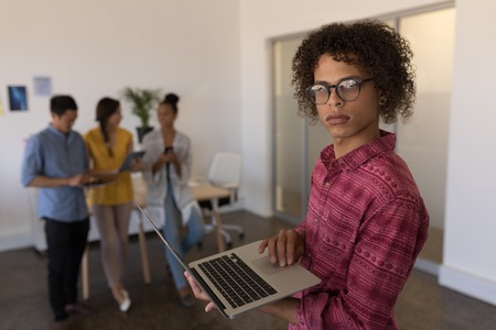 Portrait of handsome mixed race male executive using laptop in modern office with diverse colleagues interacting in the backgroundの写真素材