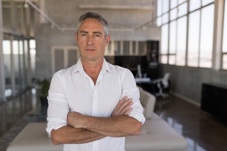 Front view of serious looking well dressed Caucasian male executive standing with arms crossed in modern officeの写真素材