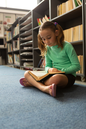Front view of Caucasian schoolgirl sitting on floor in cross-legged position and reading a book in library at elementary schoolの写真素材