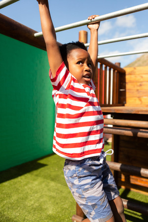 Front view of a focused cute African American schoolboy playing on horizontal ladder in the school playground on a sunny dayの写真素材