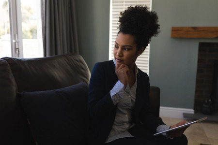 Front view of a thoughtful female real estate agent using digital tablet and looking away while sitting on a sofa in living room at homeの写真素材
