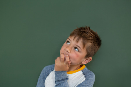 Front view of thoughtful Caucasian schoolboy standing against green chalkboard in a classroom at...