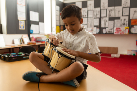 Front view of cute focused mixed-race schoolboy sitting on desk and playing bongo in a classroom at elementary schoolの写真素材